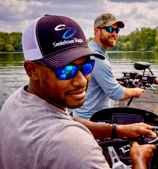 Two men on a boat in a lake, one wearing a cap and sunglasses with 'Smokehouse Bayou branding.